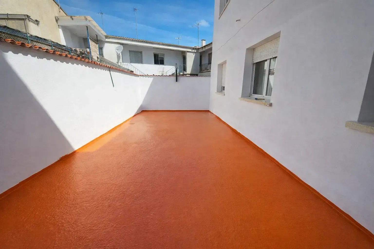 Un estrecho patio exterior con suelo texturizado de color naranja, rodeado de paredes blancas bajo un cielo azul.