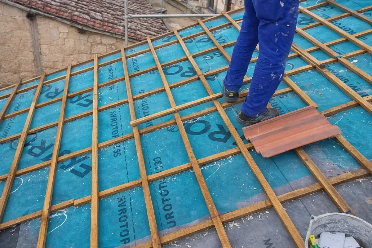 Una persona con pantalones de trabajo azules está de pie sobre la estructura de un tejado inclinado cubierto