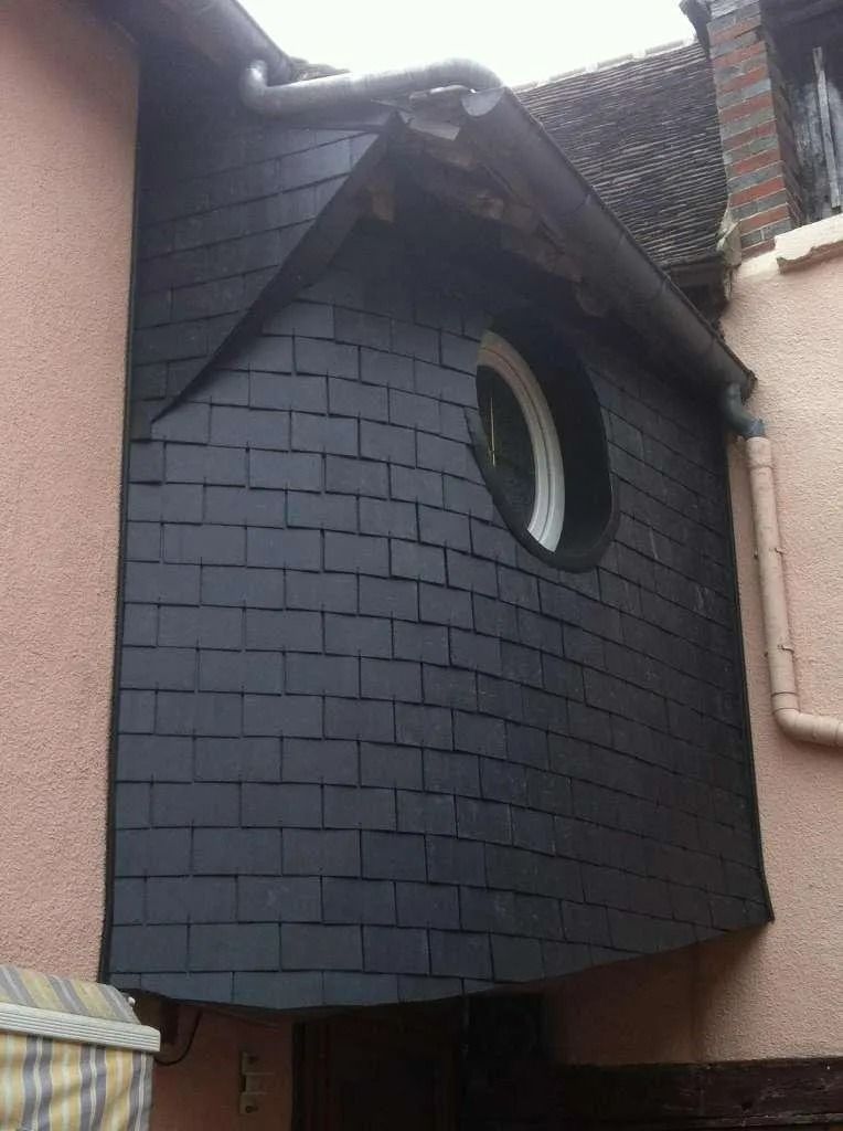 Mur extérieur recouvert d'ardoise sombre avec une fenêtre ronde, entre deux bâtiments roses, sous une gouttière.
