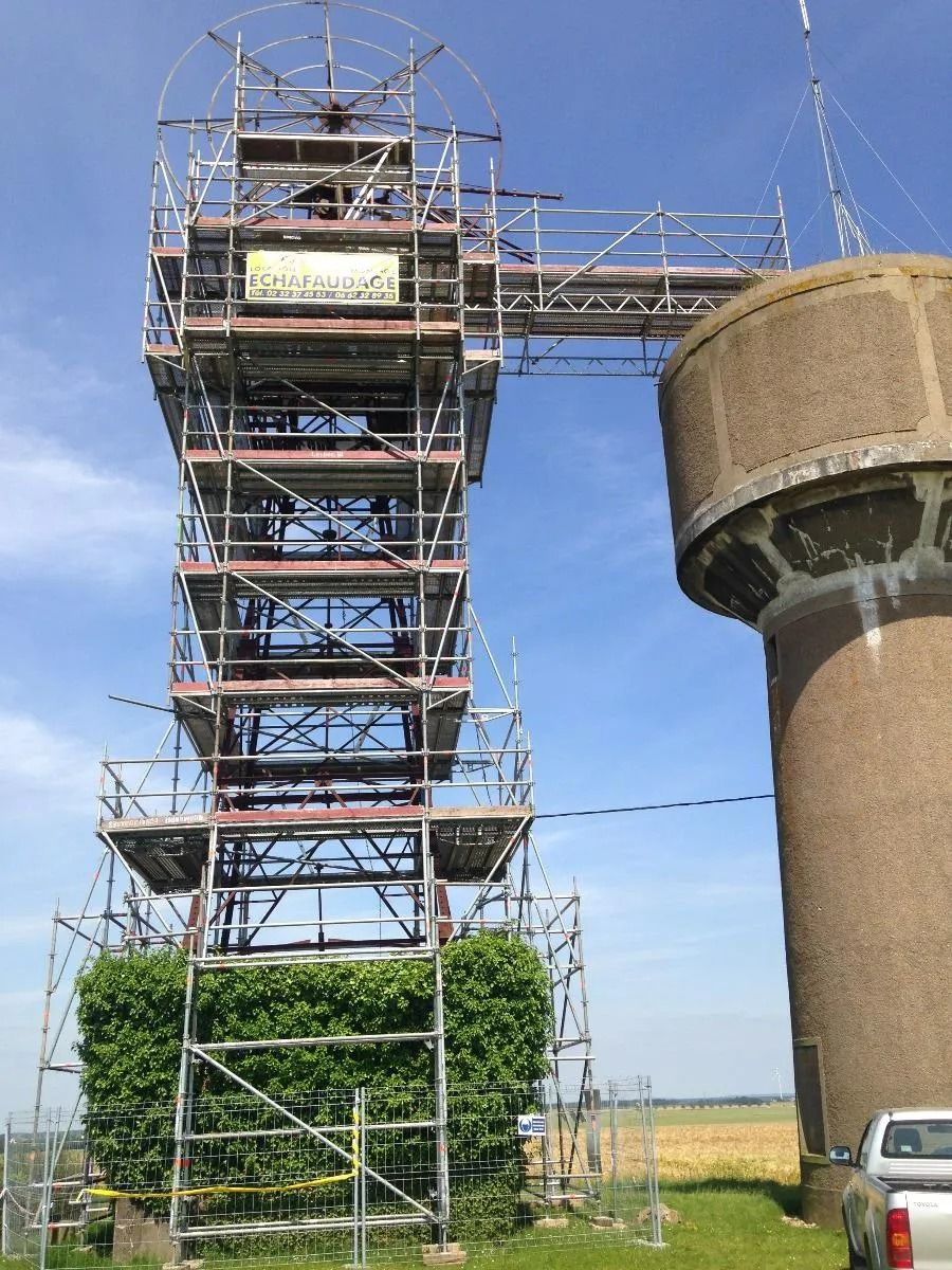 Des échafaudages entourent une haute tour recouverte de lierre, reliée par une passerelle à un château d'eau cylindrique, en plein air.