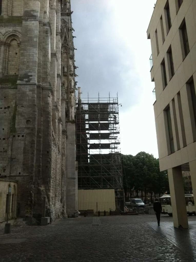 Façade de cathédrale en pierre avec échafaudages, bâtiment moderne beige, personne marchant sur des pavés.