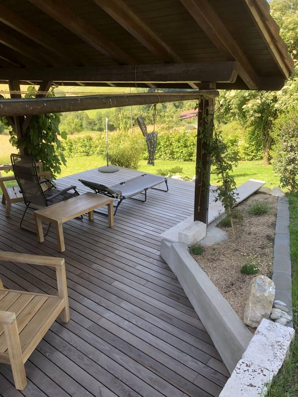 Holzterrasse mit Gartenmöbeln unter einer überdachten Veranda mit Blick auf eine grüne Landschaft.