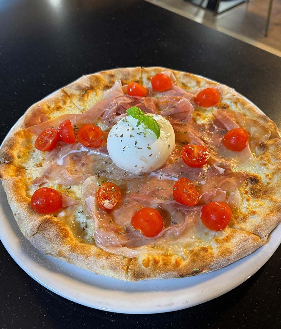 Una pizza con jamón, tomates y mozzarella en un plato blanco sobre una mesa.