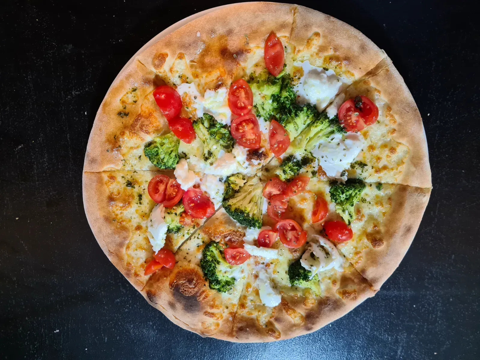 Una pizza con brócoli, tomates y mariscos sobre una mesa negra.