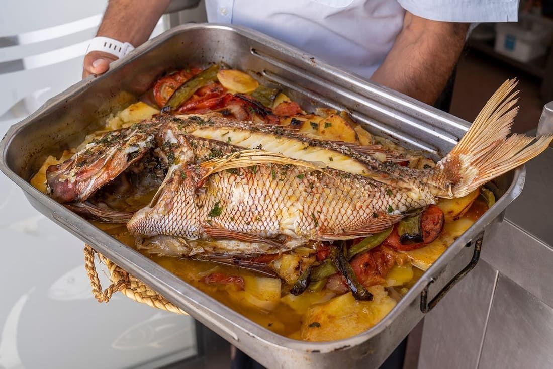 Un pescado grande asado en una bandeja de metal con verduras y rodajas de limón, sostenido por una persona en una cocina.