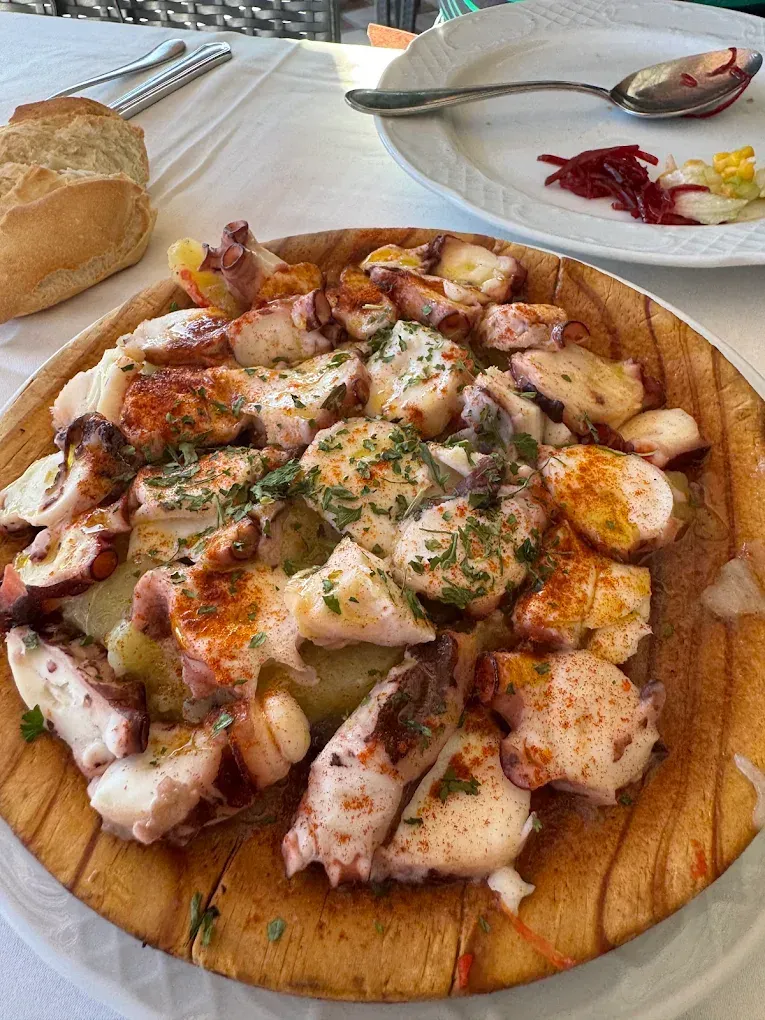 Pulpo con patatas, pimentón y perejil en plato de madera; pan, ensalada y cubiertos a la vista.