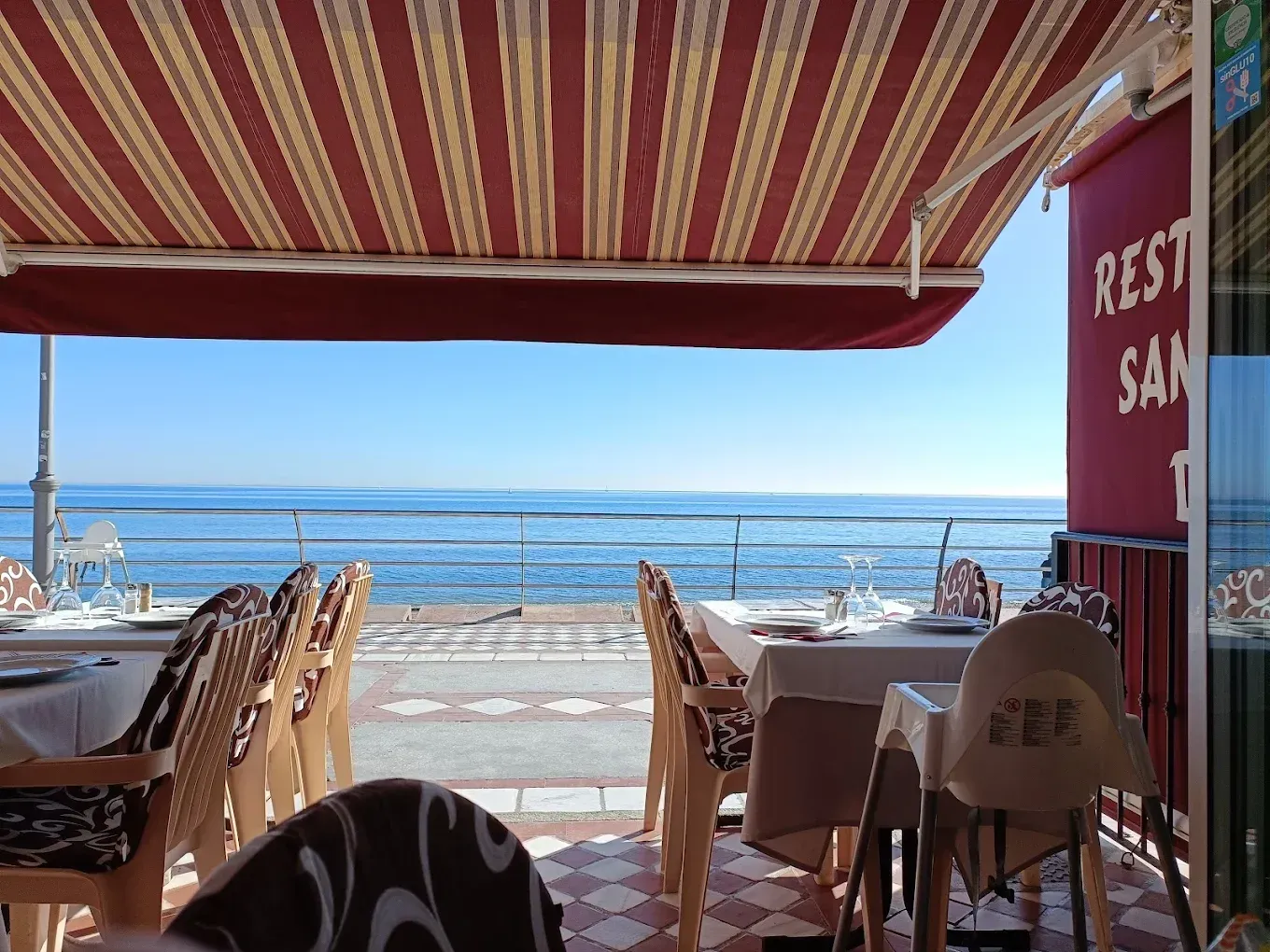 Patio del restaurante con vistas al mar, con toldo a rayas, mesas y sillas. Día soleado.
