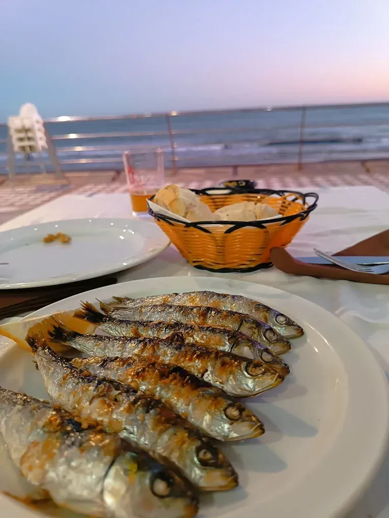 Sardinas a la parrilla en un plato, canasta de pan, vaso de jugo y vista al mar al atardecer.