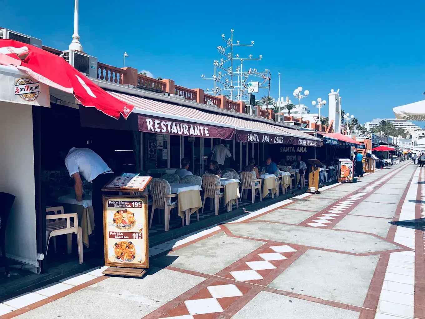Restaurantes con toldos rojos bordean un soleado paseo marítimo. La gente cena al aire libre.
