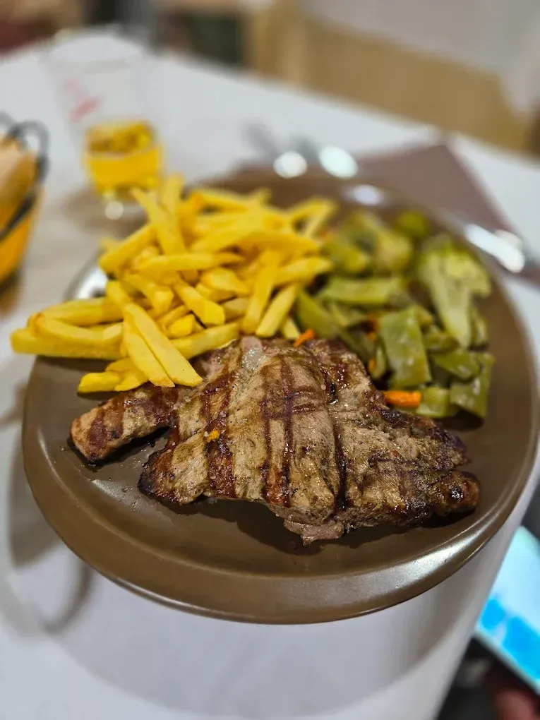 Bistec a la parrilla con patatas fritas y judías verdes en un plato marrón, sobre un mantel blanco.