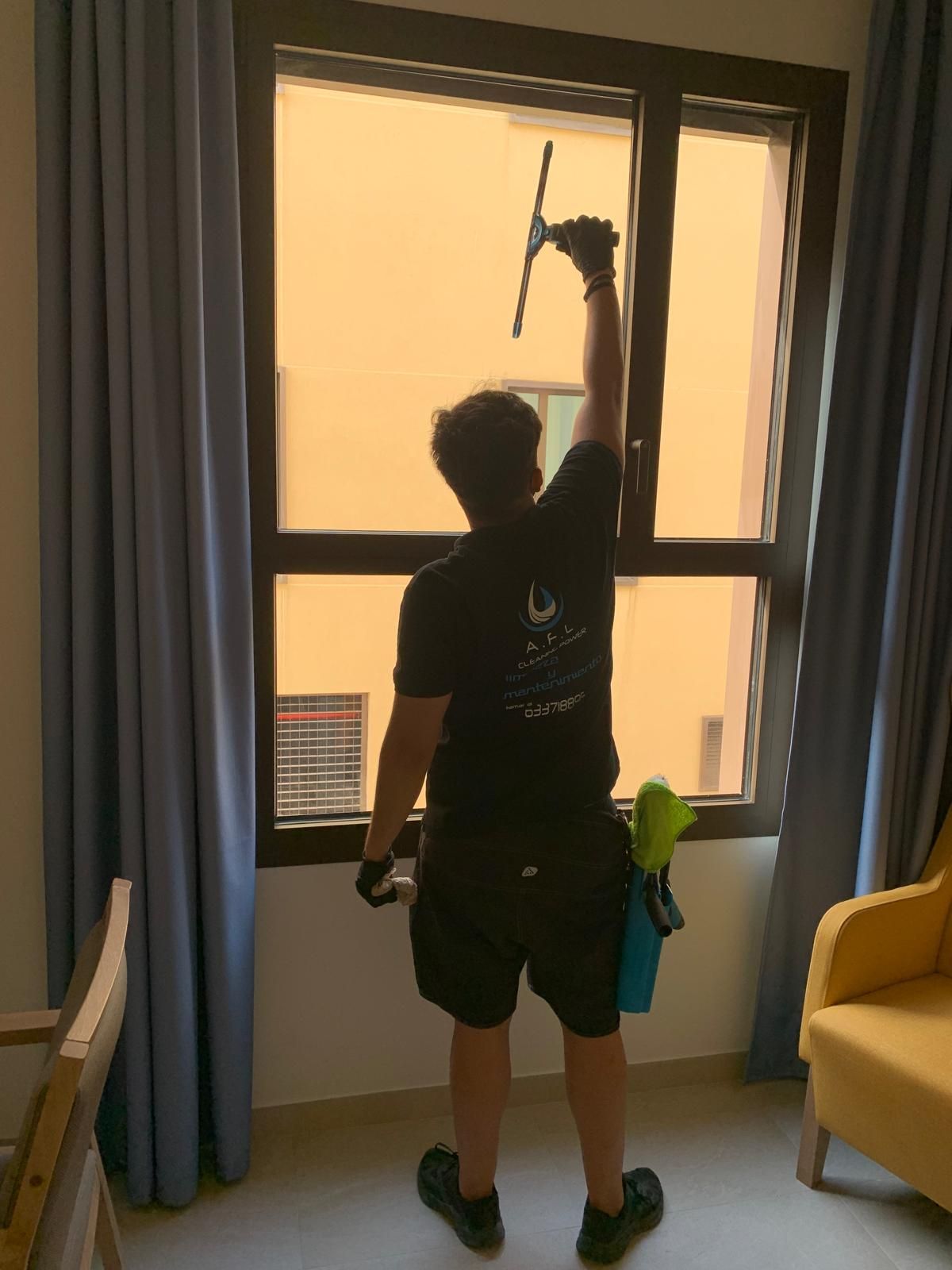 Persona limpiando una ventana con una rasqueta, vista desde atrás. Interior, cortinas y silla amarilla a la vista.