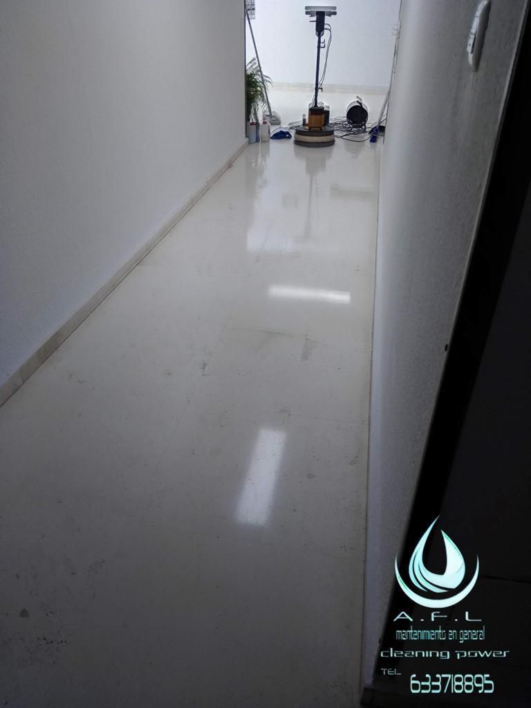 Pasillo largo con suelo pulido de color claro, equipo de limpieza al fondo. Paredes blancas, puerta oscura.