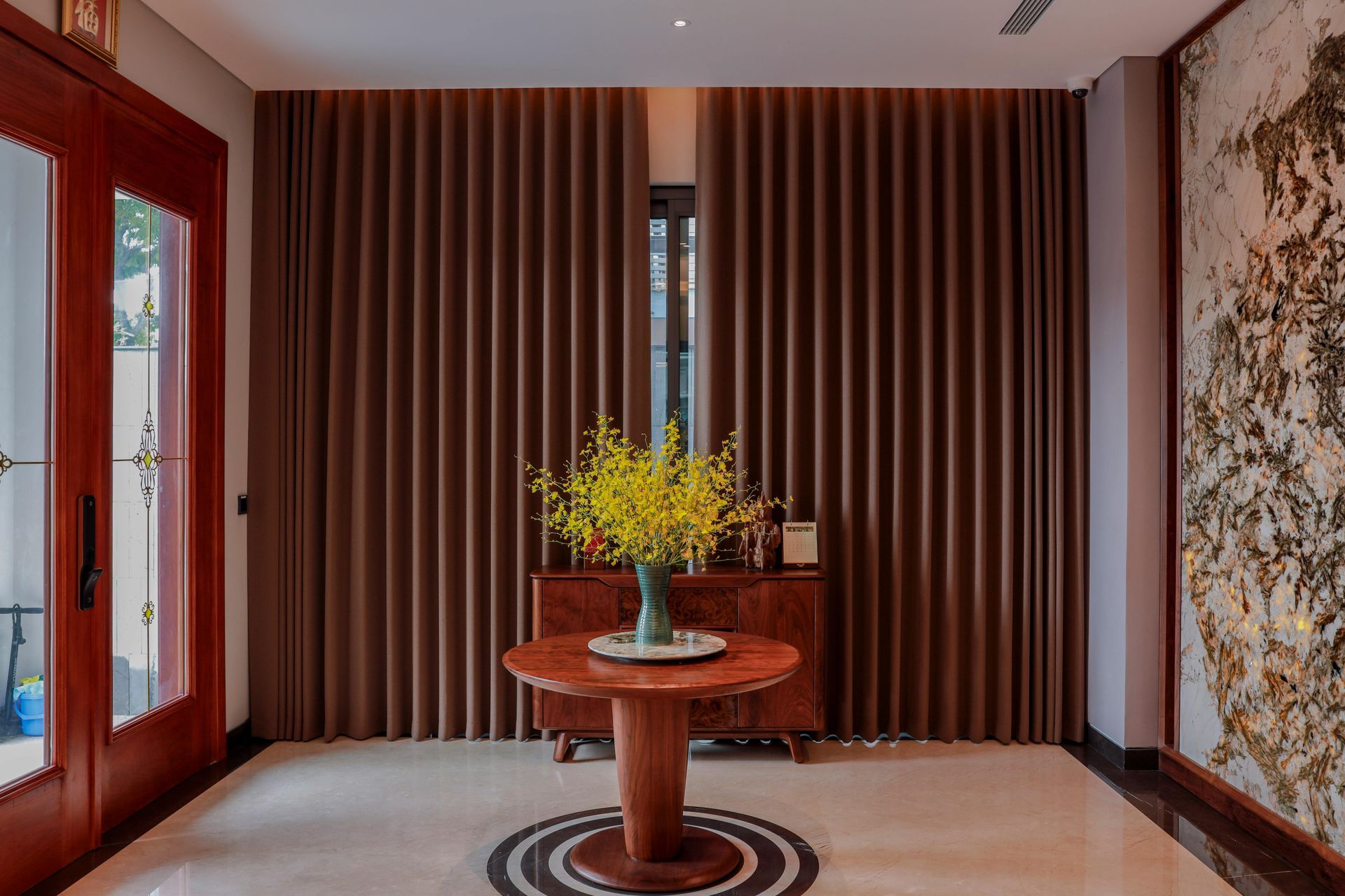 Recibidor con cortinas marrones, una mesa de madera, un arreglo floral y una pared decorativa.