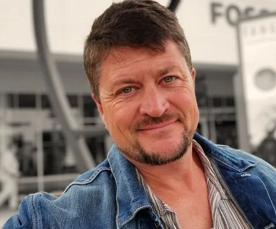 Man smiling in denim jacket outdoors, blurred background, day.