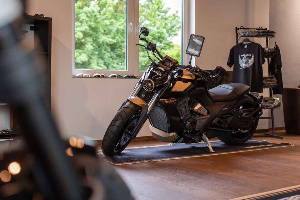 Ein schwarzes Motorrad steht in einem Innenraum in der Nähe eines Fensters; im Hintergrund ist ein Kleiderständer zu sehen.