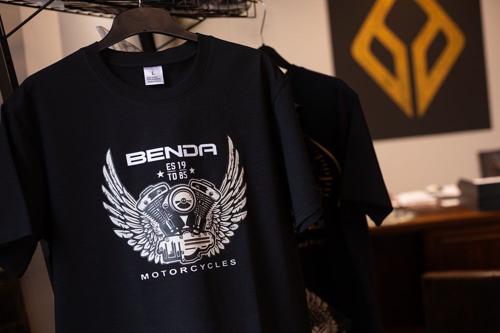 Ein schwarzes Benda Motorrad-T-Shirt hängt in einem Geschäft oder Ausstellungsraum an einem Kleiderständer.