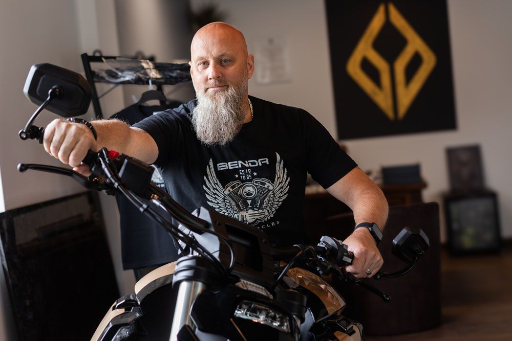 Bärtiger Mann auf einem schwarzen Motorrad in einem Innenraum, im Hintergrund ein schwarz-goldenes Wandlogo.