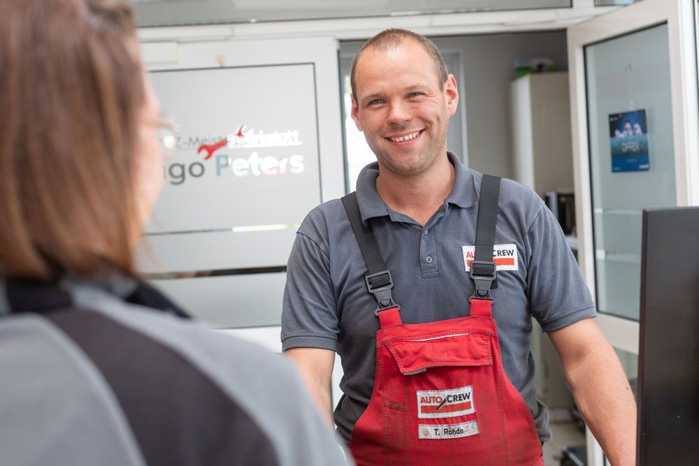 Ein lächelnder Angestellter in rotem Overall steht an einem Bedienschalter und unterhält sich im Vordergrund mit einem Kunden.