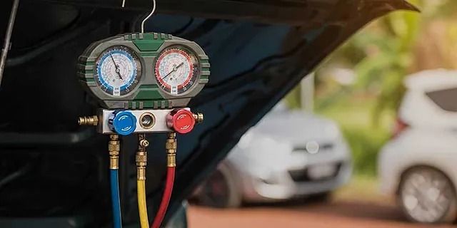 Nahaufnahme der Manometer der Klimaanlage eines Autos mit blauen, gelben und roten Schläuchen unter einer geöffneten Motorhaube.