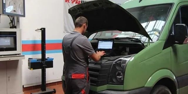Ein Mechaniker untersucht in einer Werkstatt einen grünen Lieferwagen mit geöffneter Motorhaube.