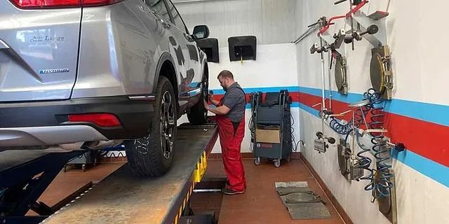 Ein Mechaniker in rotem Overall inspiziert einen silbernen SUV auf einer Hebebühne in einer Autowerkstatt.