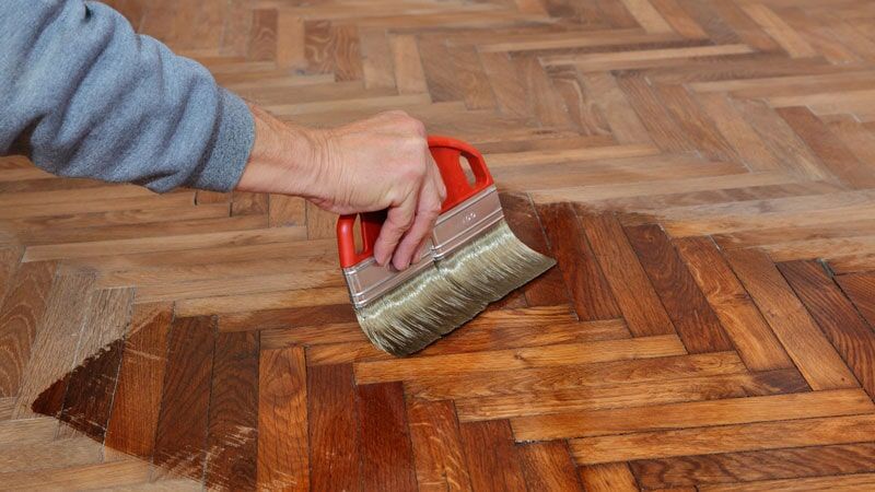 Pessoa aplicando acabamento em um piso de parquet de madeira com um pincel.