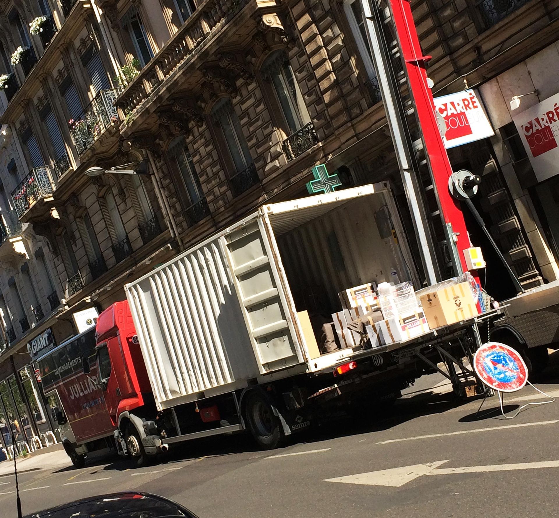 Vue inclinée d'un camion rouge transportant un conteneur maritime dans une rue de la ville, à proximité de bâtiments et de panneaux de signalisation.