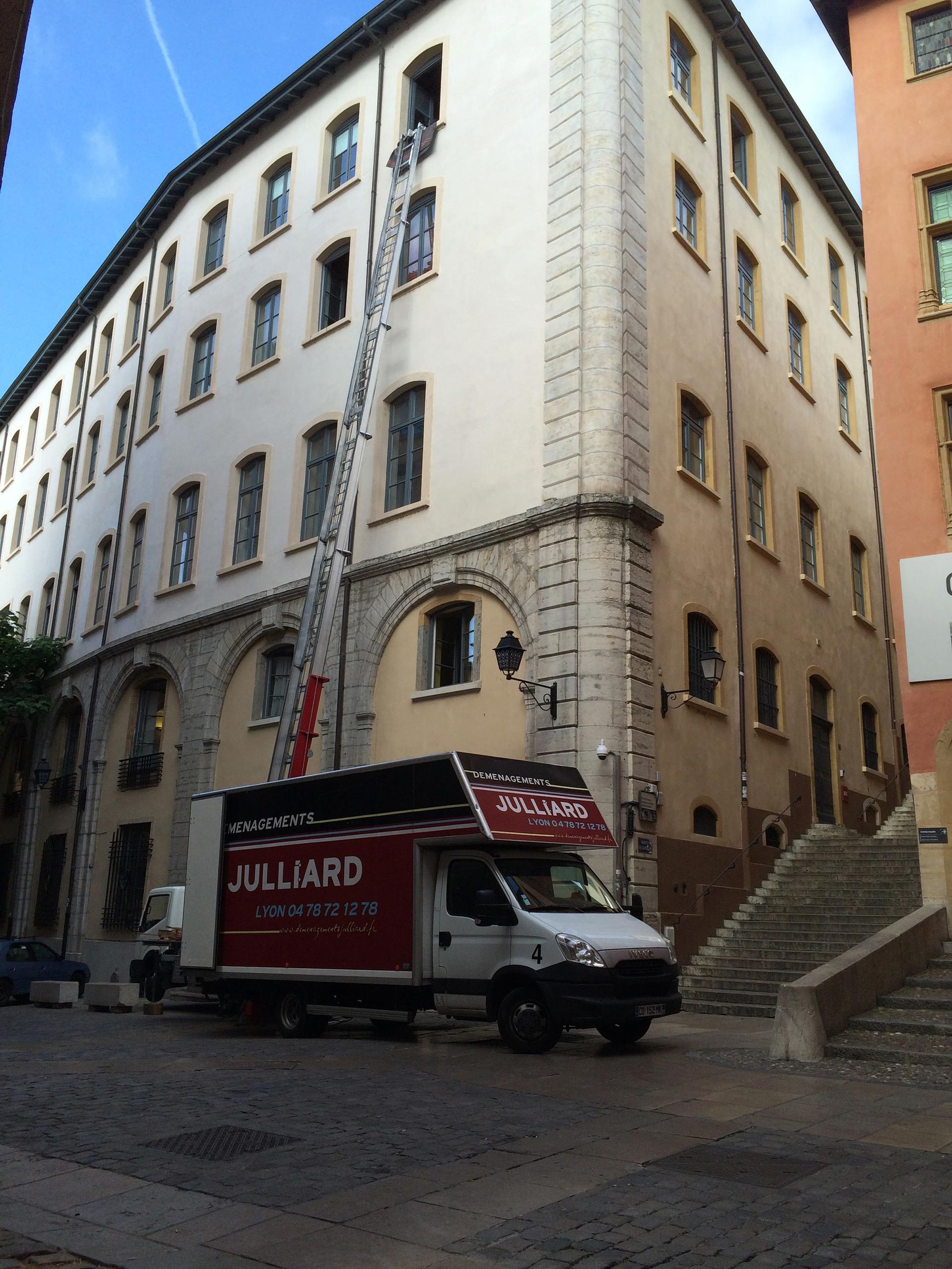 Bâtiment blanc d'angle dans une rue pavée, avec une camionnette de livraison rouge garée devant.
