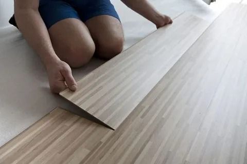 Persona arrodillada, instalando tablones de piso de madera de color claro sobre una base blanca.