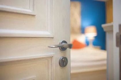 Puerta blanca entreabierta, revelando un dormitorio con una cama, una lámpara y una pared azul.