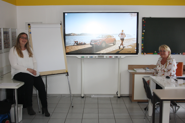 Zwei Frauen sitzen vor einem großen Bildschirm mit dem Bild eines Autos darauf
