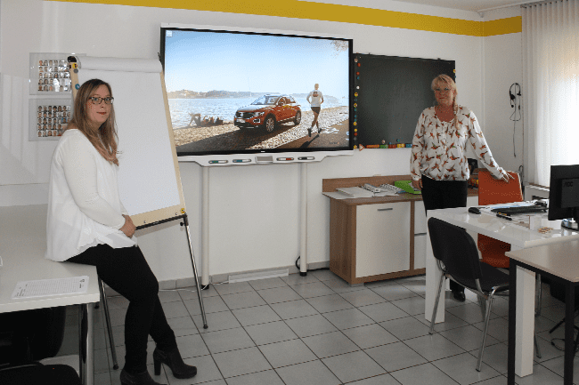 Zwei Frauen stehen vor einem großen Bildschirm mit dem Bild eines Lastwagens darauf