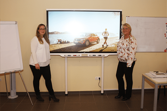Zwei Frauen stehen vor einem großen Bildschirm mit dem Bild eines Autos darauf