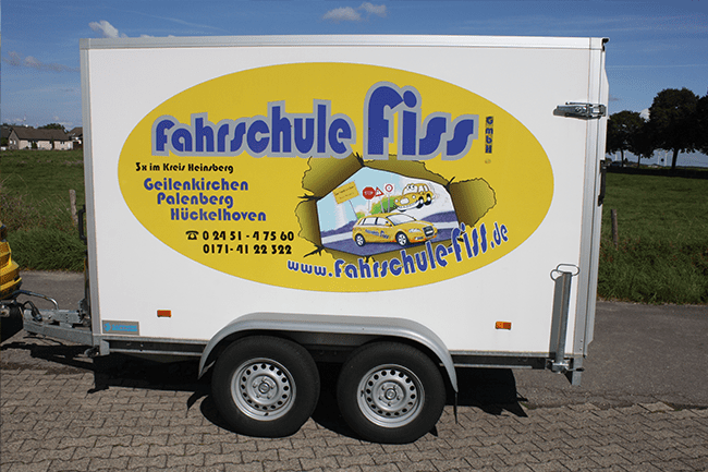 Ein weißer Anhänger mit einem gelben Aufkleber mit der Aufschrift Fahrschule Fissi