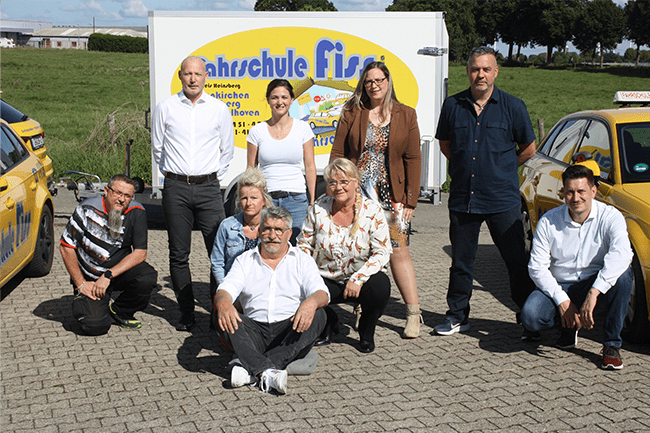 Eine Gruppe von Menschen posiert für ein Foto vor einem Anhänger mit der Aufschrift „Fahrschule Fiss“