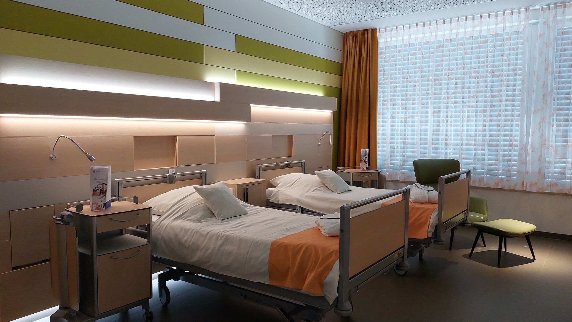 Ein Krankenhauszimmer mit zwei Betten, einem großen Fenster und bunten Wandpaneelen. Neben den Betten stehen Nachttische.