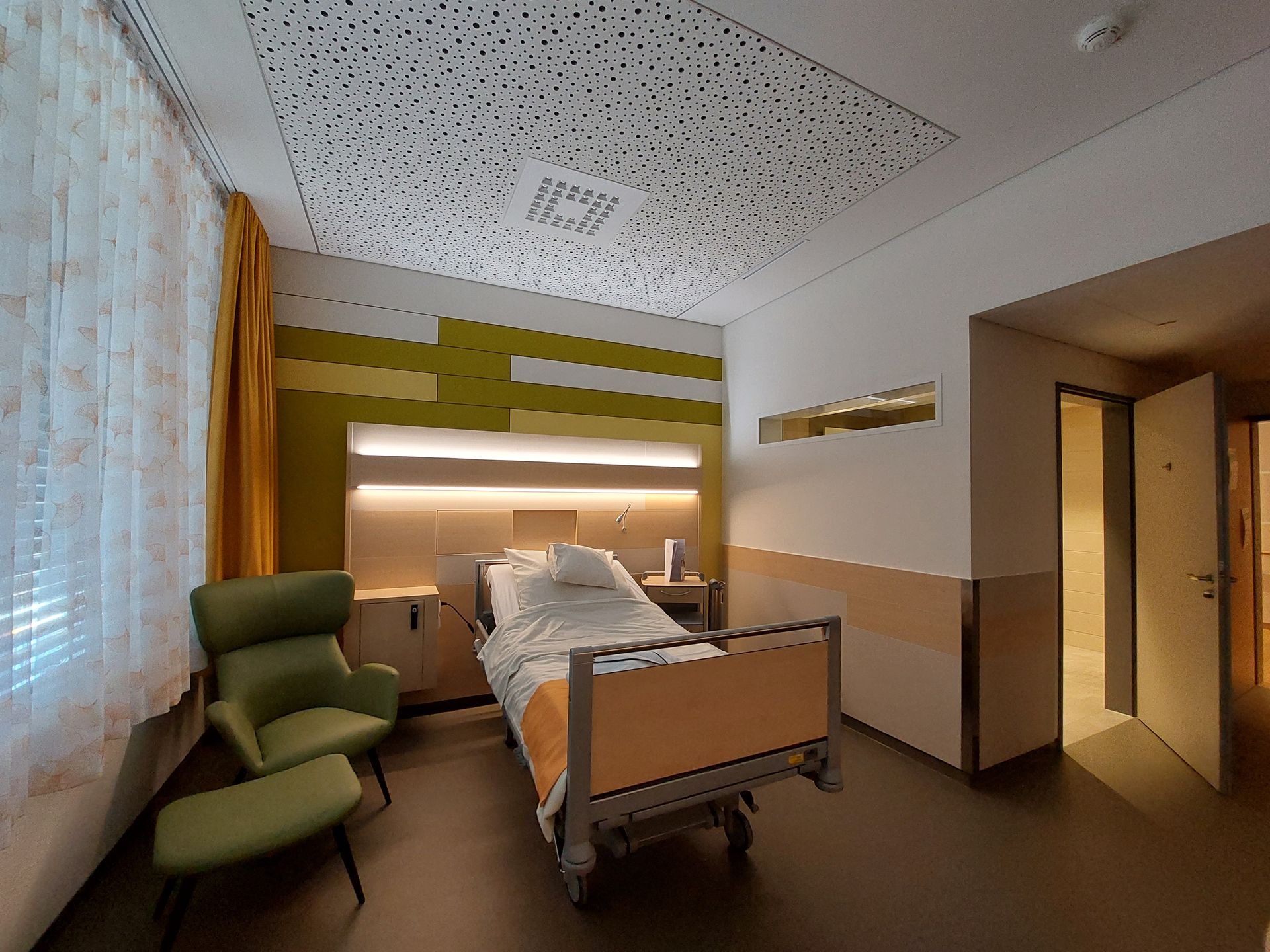 Ein Krankenhauszimmer mit Bett, Sessel und Ottomane.