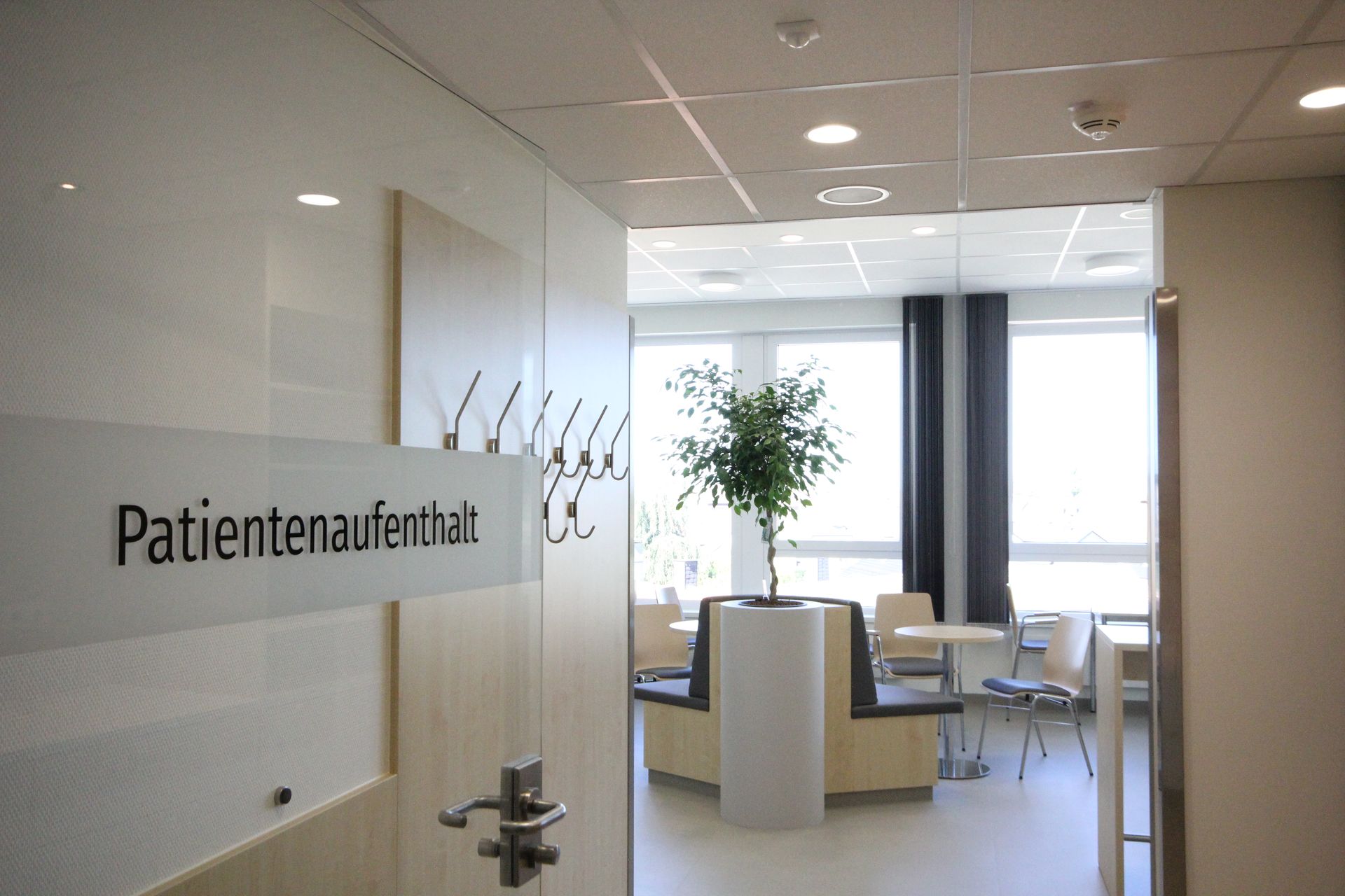 Eine Glastür mit dem Schriftzug „Patientenaufenthalt“ führt in einen hellen Wartebereich.