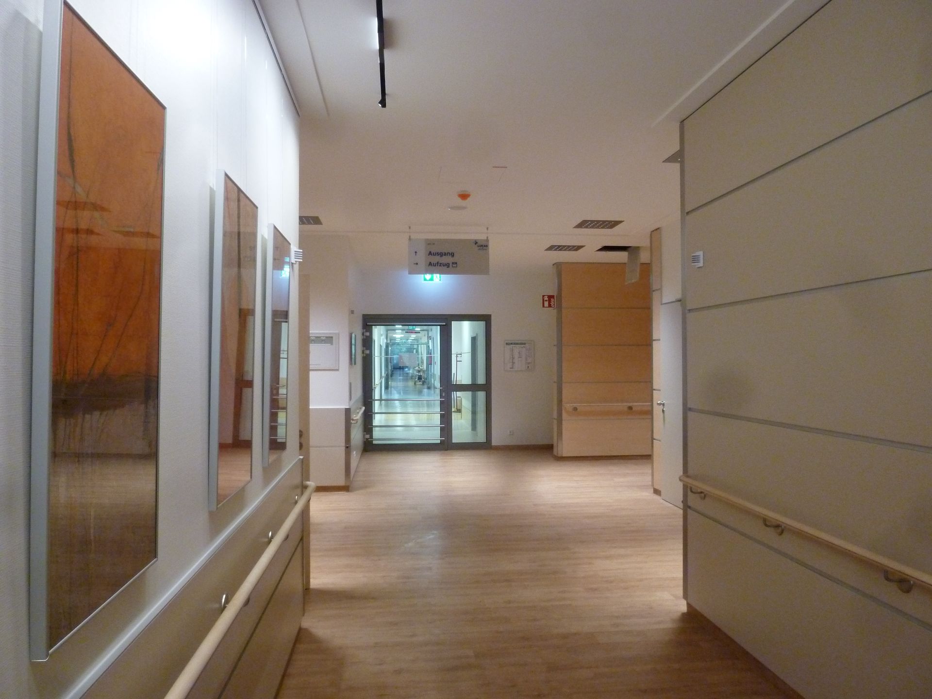 Langer Flur in einem Krankenhaus mit Holzböden, hellen Wänden und Kunstwerken. Eine Tür führt nach draußen.