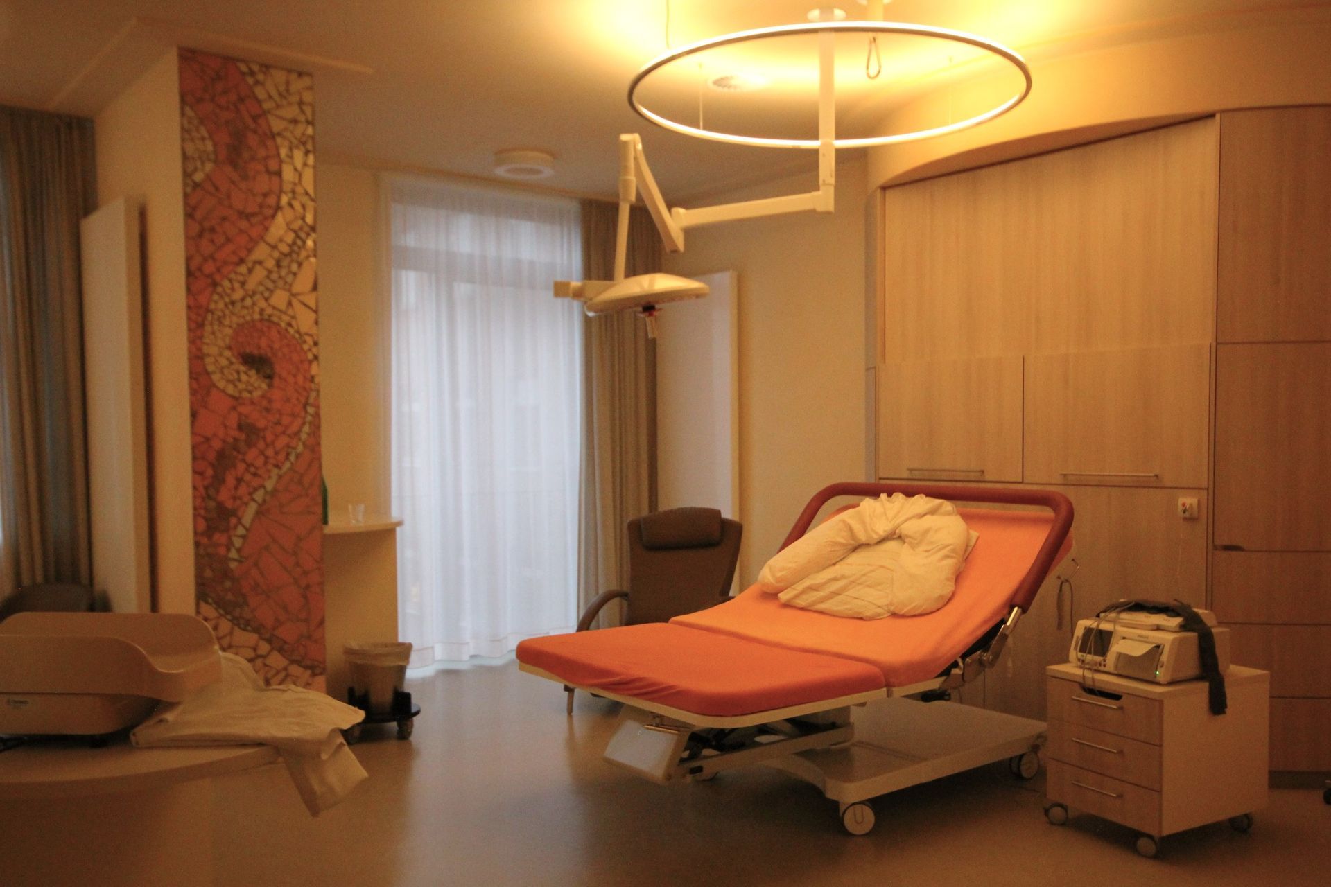 Ein Kreißsaal mit Bett, medizinischer Ausstattung und dekorativer Wand. Der Raum ist mit warmem Licht beleuchtet.