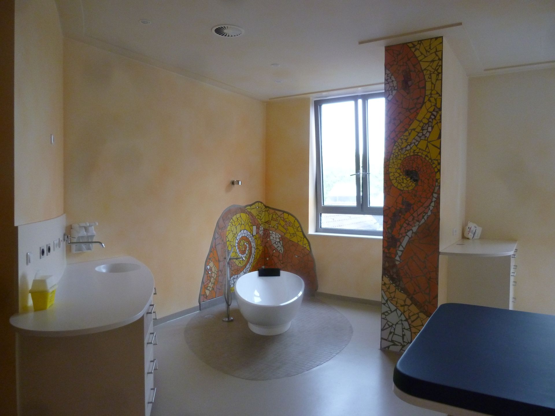 Ein barrierefreies Badezimmer mit mosaikverzierter Wand, Toilette, Waschbecken und Fenster.