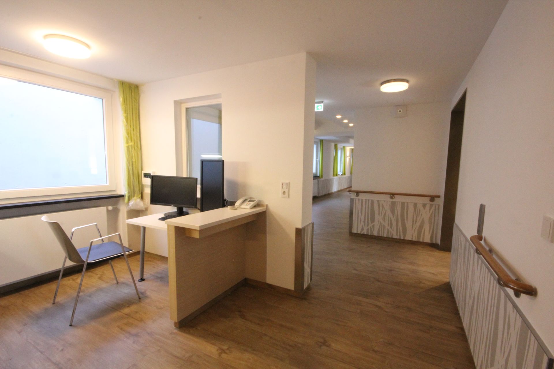 Ein Büroempfangsbereich mit Schreibtisch, Stuhl und Computer, der zu einem Flur mit Geländer führt. Helle Holzböden.