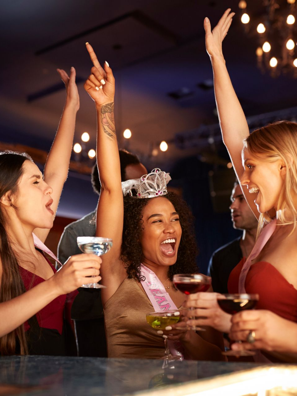 Des personnes font la fête dans un bar, les bras levés et un verre à la main. La femme au centre porte un diadème et une écharpe.