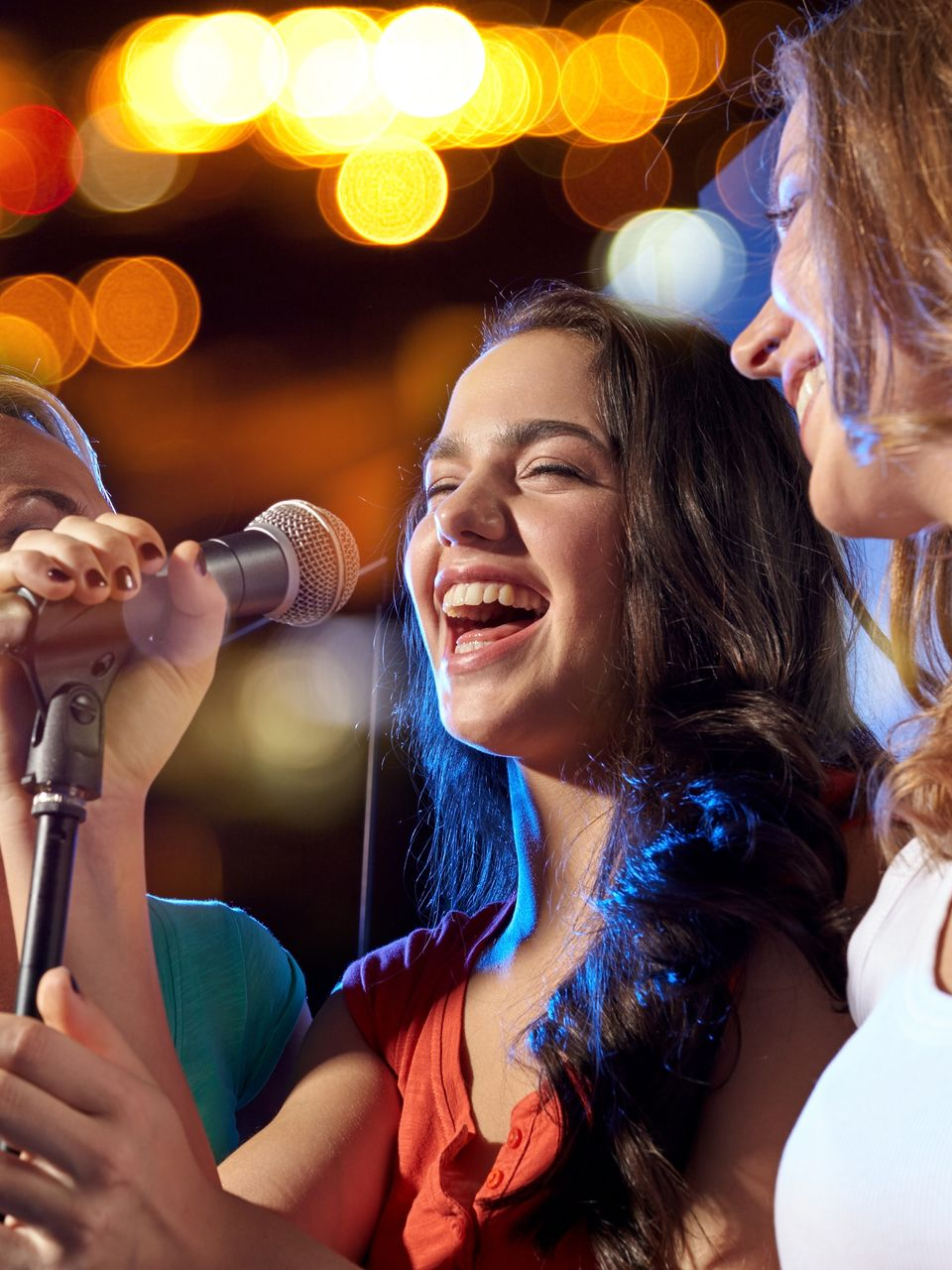 Une femme chante dans un micro, en riant, avec des amis, la nuit. Lumières vives en arrière-plan.