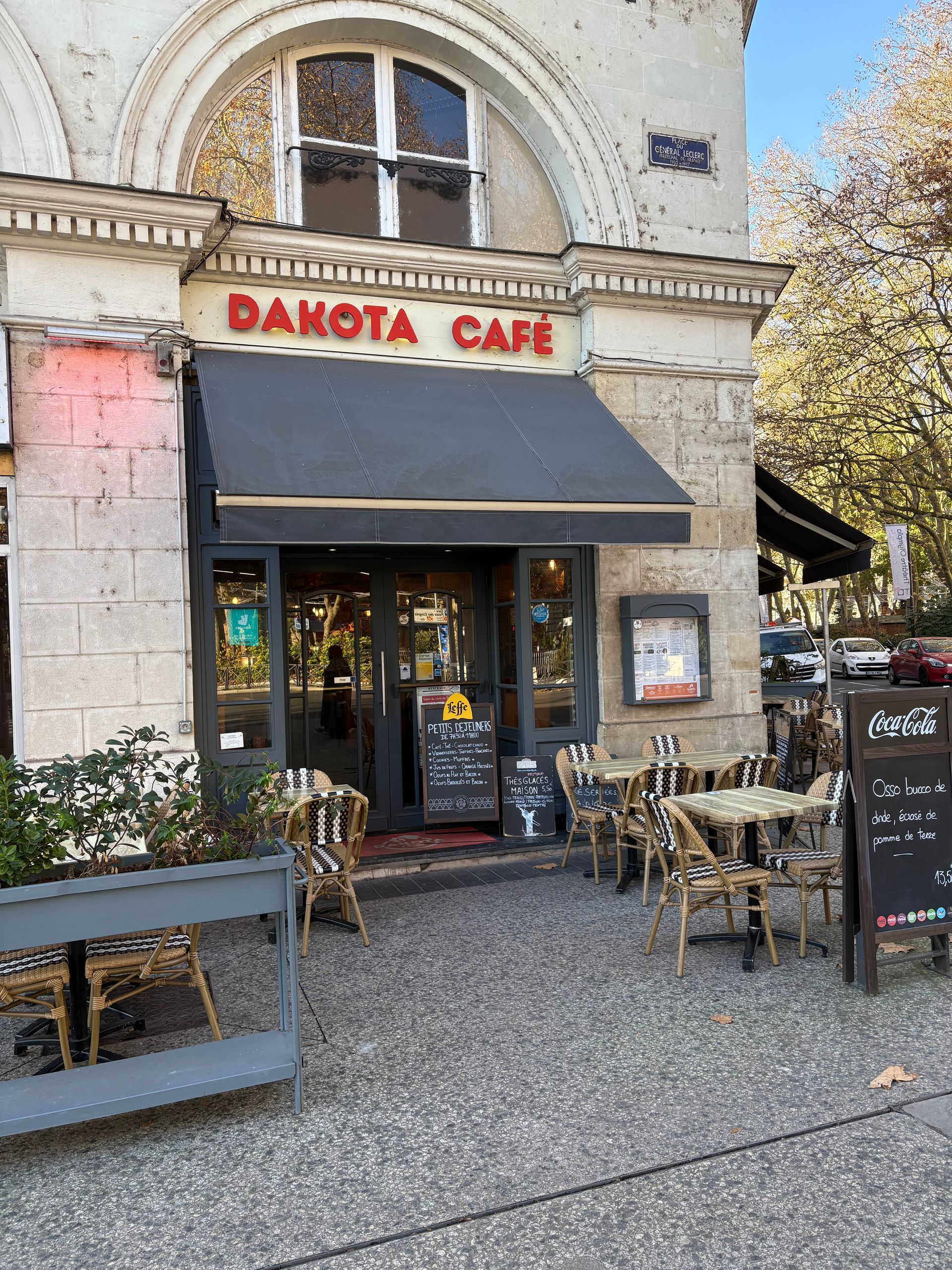 Entrée du Dakota Café avec terrasse. Enseigne rouge sur un auvent noir, façade en pierre.