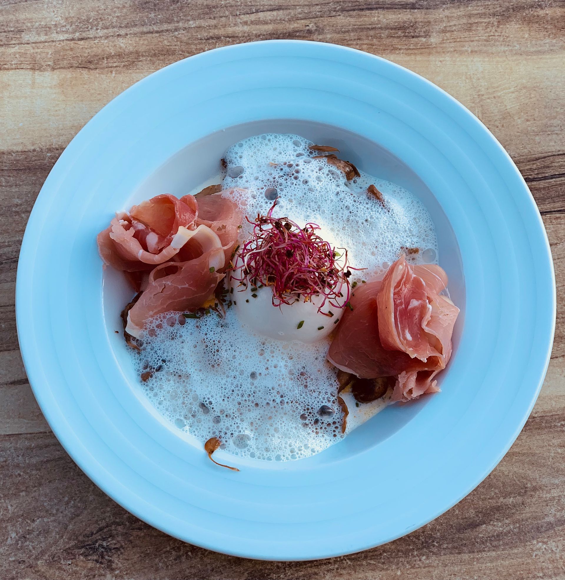 Un plat d'œuf poché avec mousse, prosciutto et micro-pousses rouges sur une assiette bleu clair.