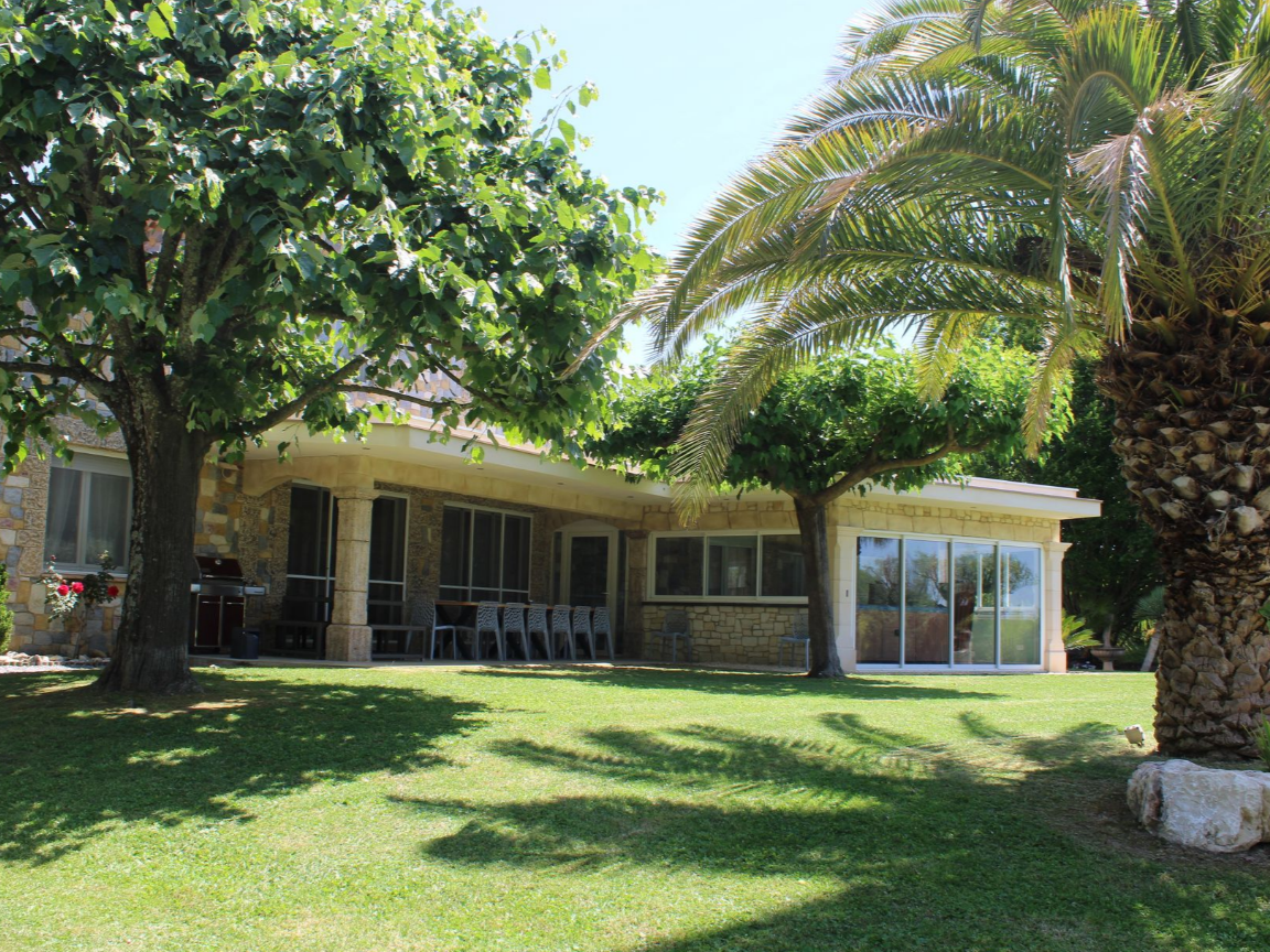Maison en pierre avec porche, pelouse et arbres, dont un palmier au premier plan.