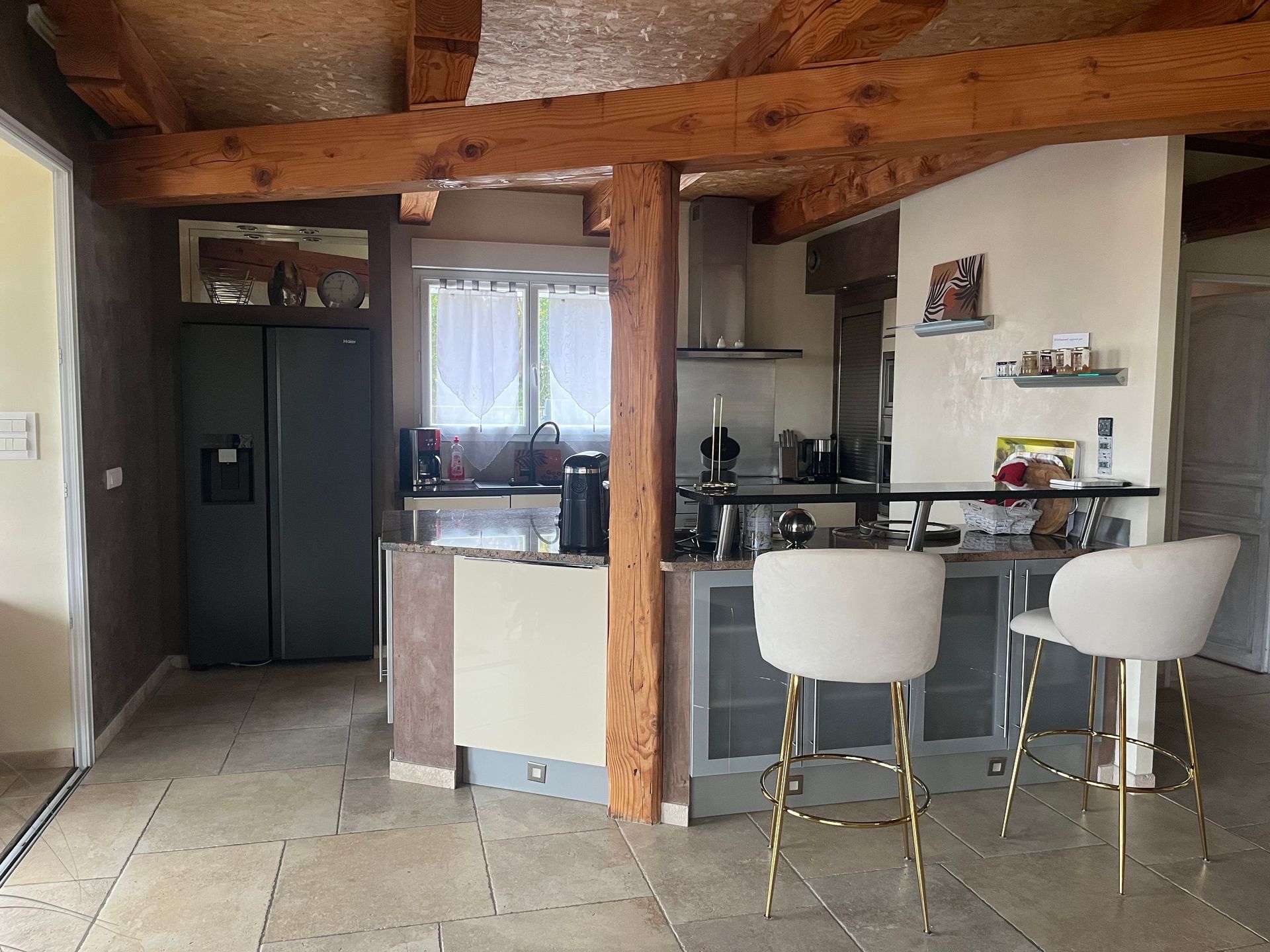 Cuisine avec réfrigérateur, comptoirs en granit et poutres apparentes. Deux tabourets de bar noirs sont disposés au comptoir.