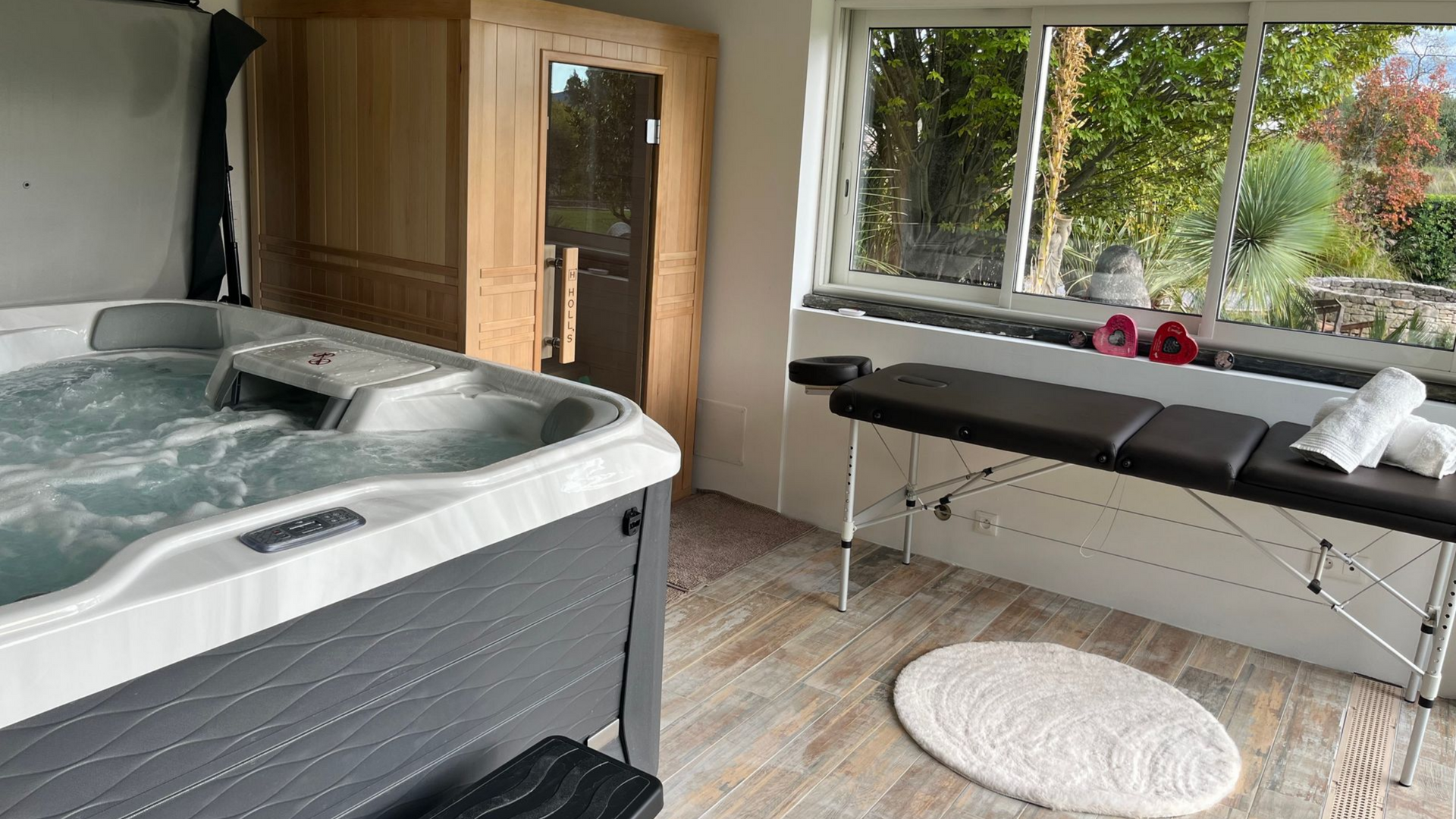 Bain à remous, sauna et table de massage dans une pièce avec une fenêtre donnant sur un jardin.