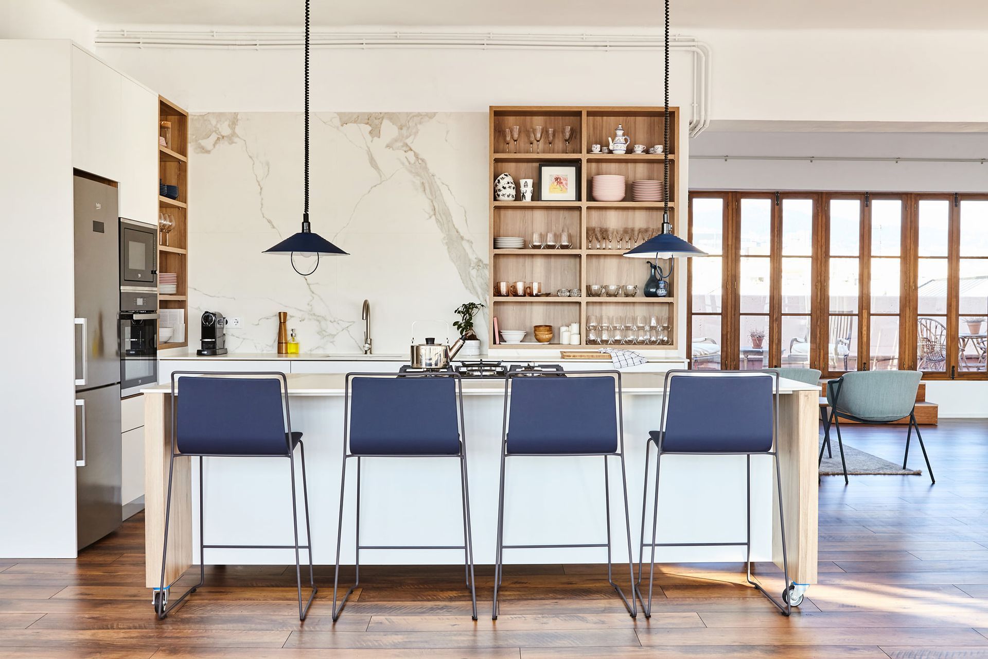 Cocina moderna con isla, cuatro taburetes de bar azules, luces colgantes, estantes de madera, protector contra salpicaduras de mármol y grandes ventanales.
